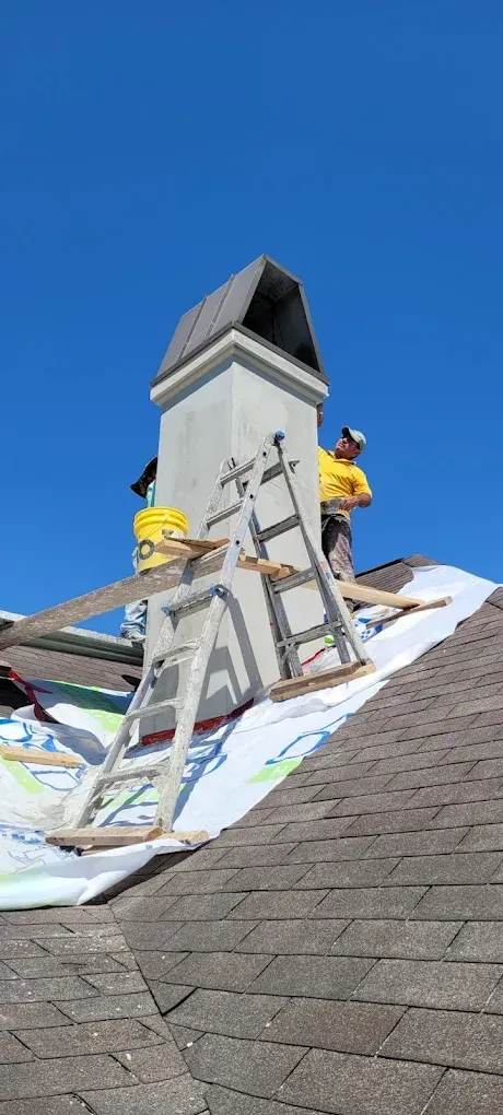 Two workers on a roof, near a chimney. One is on the chimney with a ladder. Bright blue sky overhead.