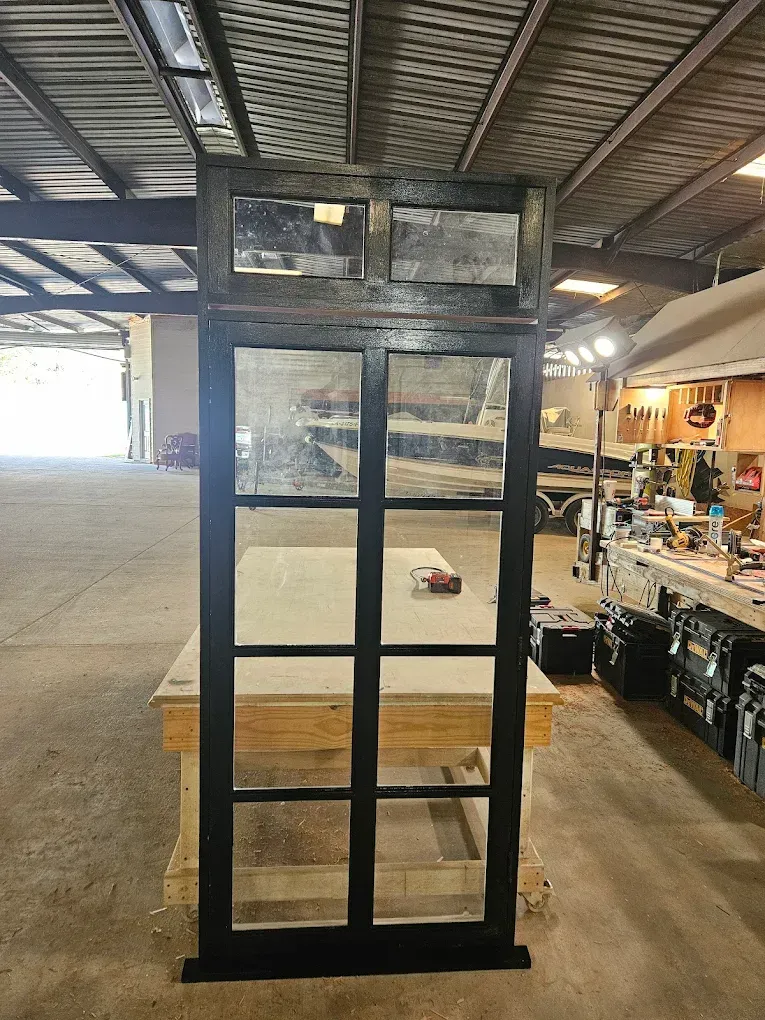 Black-framed window with six panes. It rests on a wooden work surface inside a workshop.