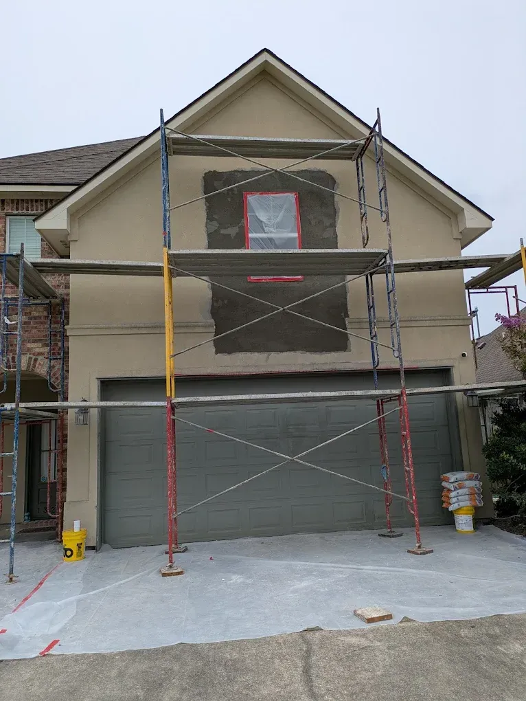 House exterior with stucco repair in progress, scaffolding around a window, garage door closed.
