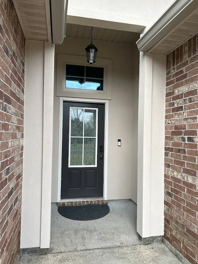 Entryway with dark brown door, small window above, brick and tan walls, black door mat.
