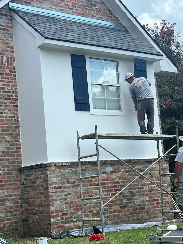 Man painting the exterior of a house, standing on a scaffold near a window with blue shutters.