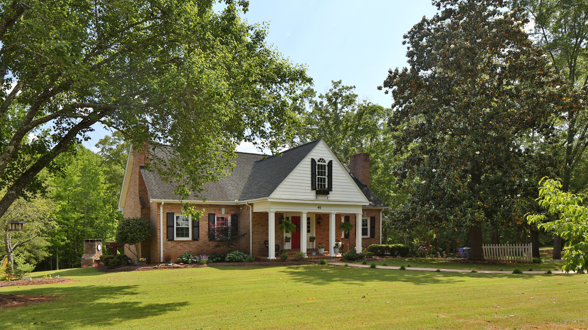 Brick house with white porch and shutters, surrounded by green trees on a grassy lawn.