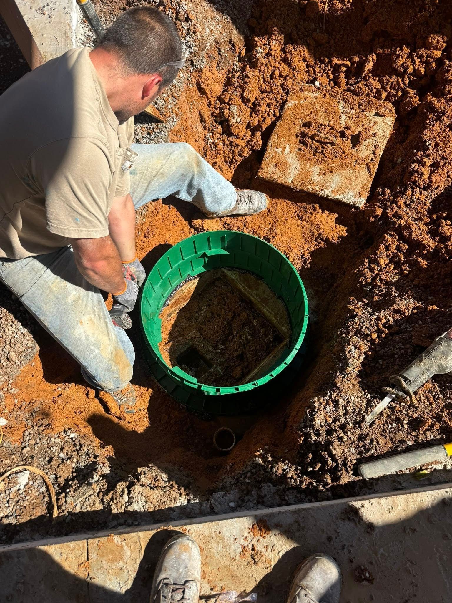 Worker kneeling in a dirt pit beside a green bucket during excavation work
