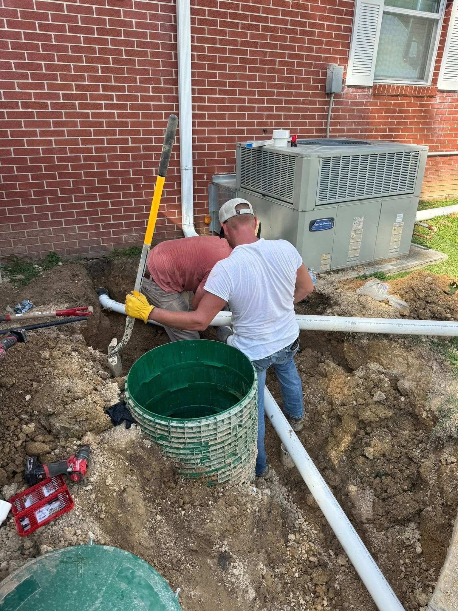 Worker in white shirt crouches by a green pit near pipes and a generator beside a brick wall.