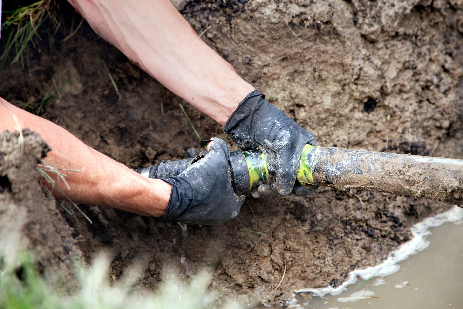 Gloved hands pulling a muddy pipe from wet ground during excavation