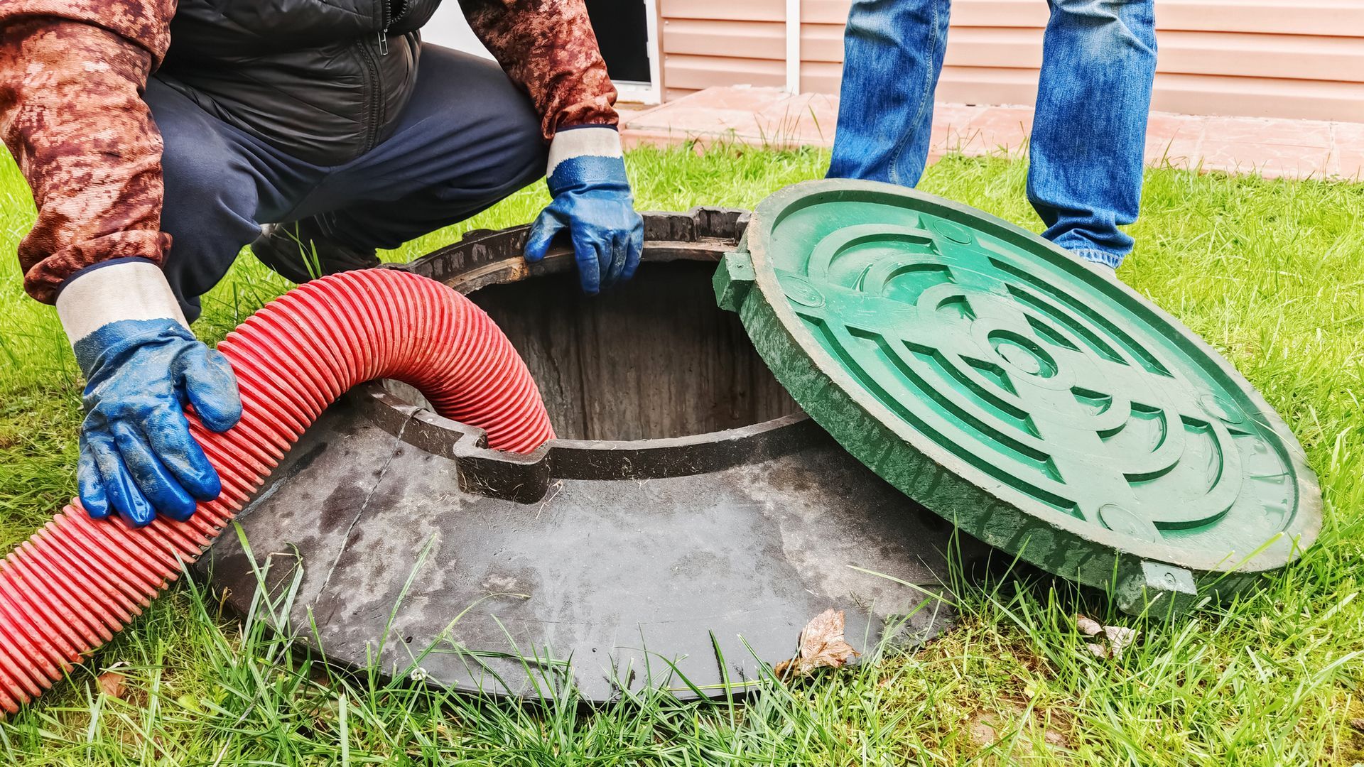 Workers in gloves removing a manhole cover from a grassy lawn with a red corrugated hose inserted.