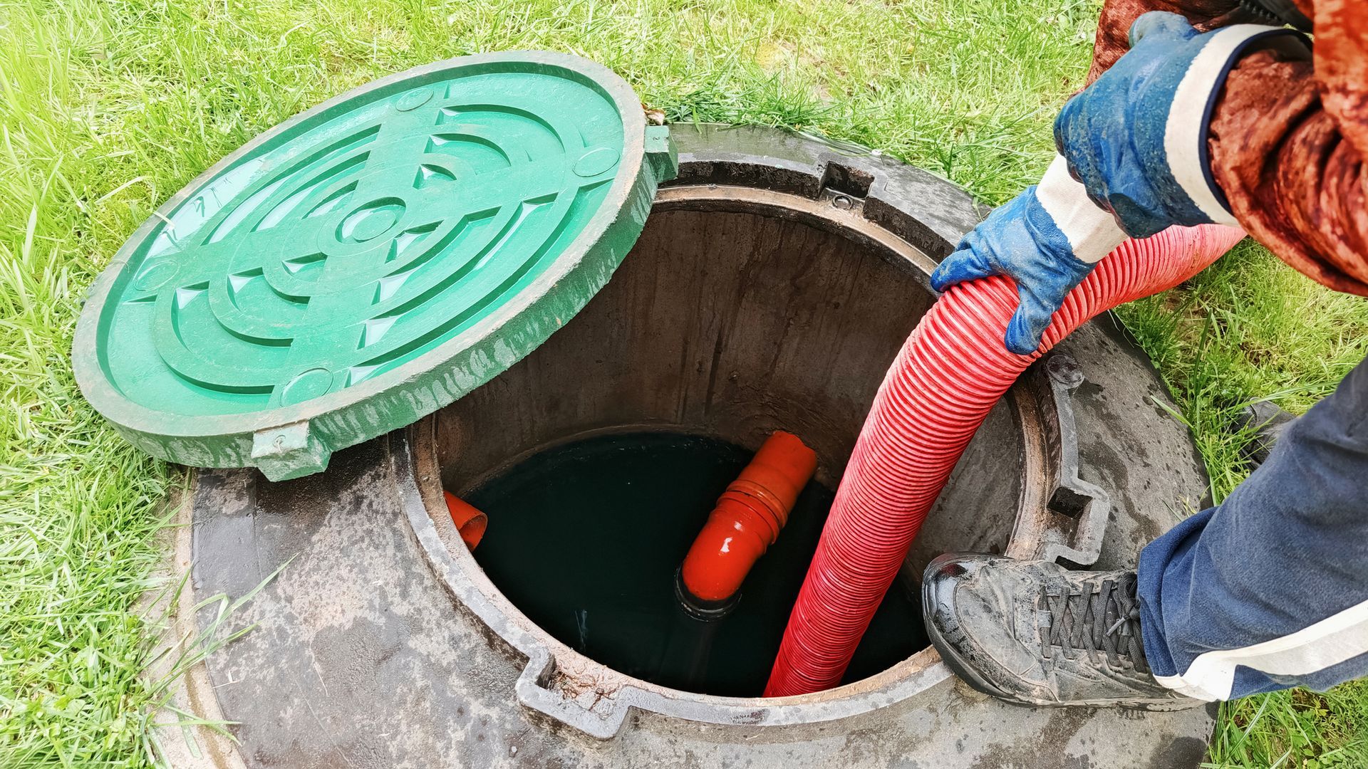 Worker uses red hose to pump water from an open manhole with green lid nearby.