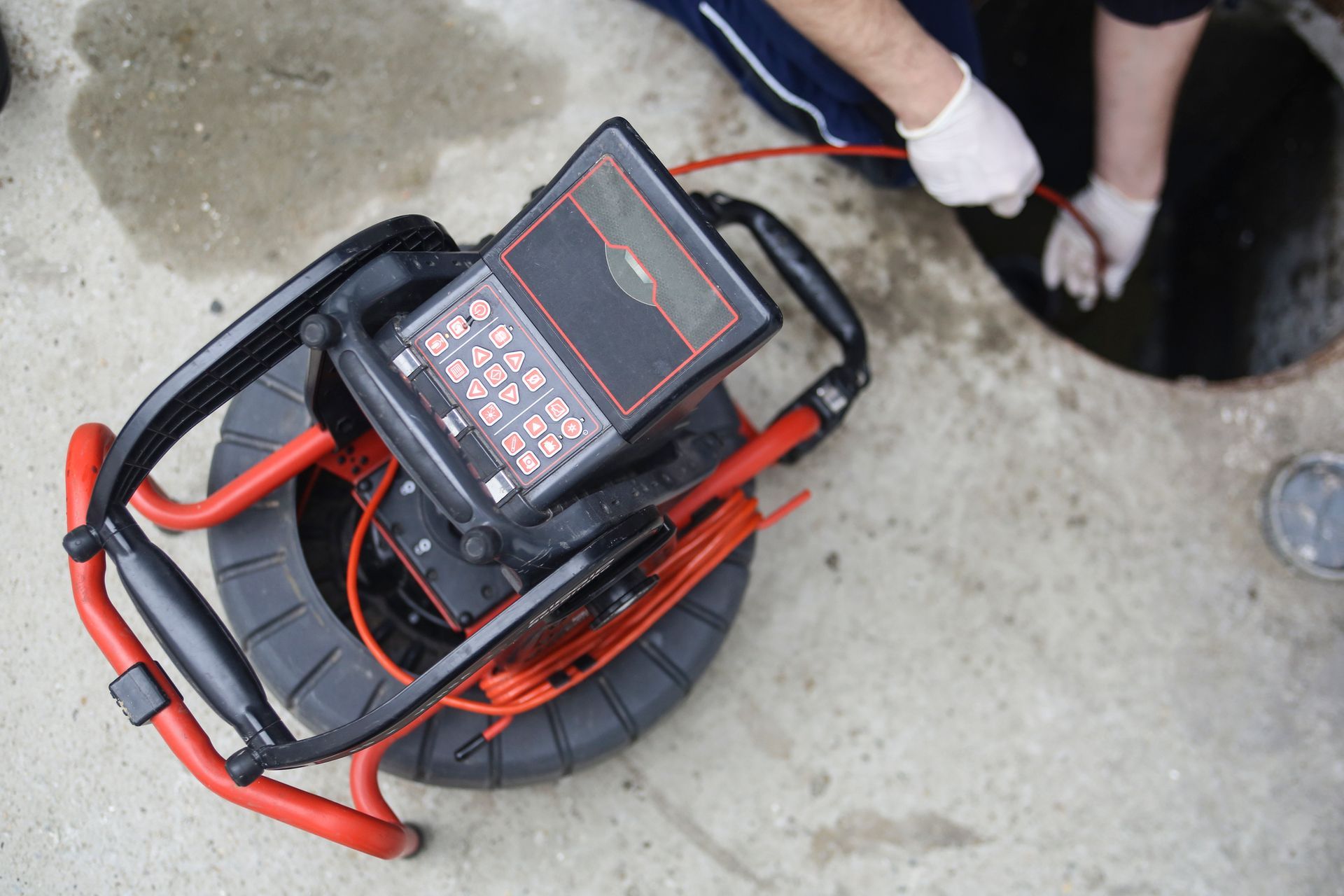 Portable cable reel with a black-and-red inspection device on top, on a concrete floor near a person’s feet
