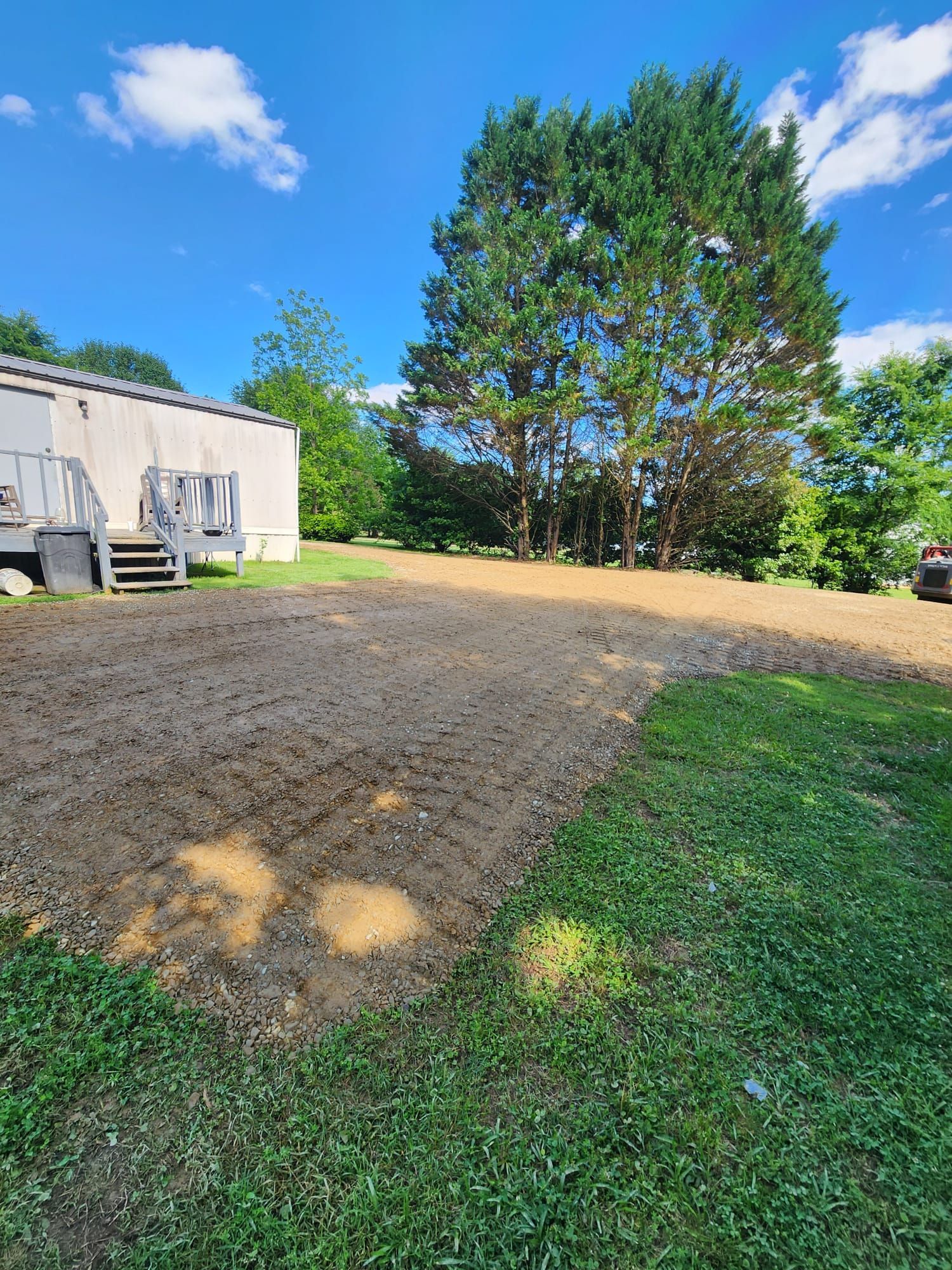 Gravel driveway beside a light-colored house, bordered by green grass and tall trees under a bright blue sky.