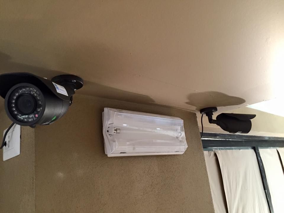 Due telecamere di sicurezza nere montate su un soffitto e una parete beige, con una lampada fluorescente.