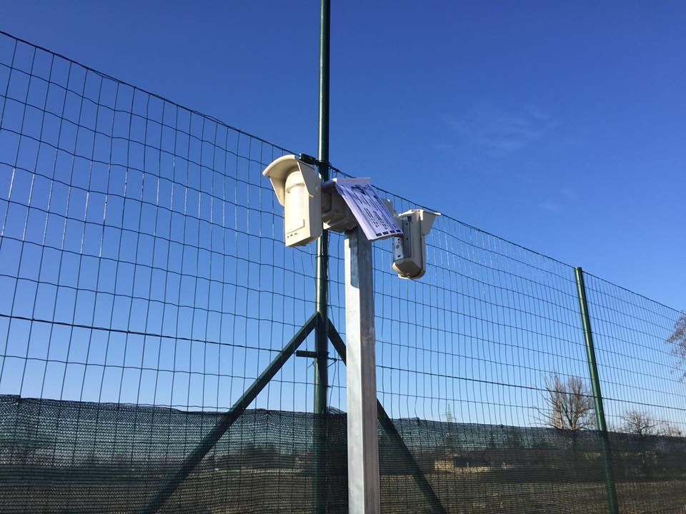 Stazione meteorologica montata su un palo con un pannello solare, dietro una recinzione metallica sotto un cielo azzurro.