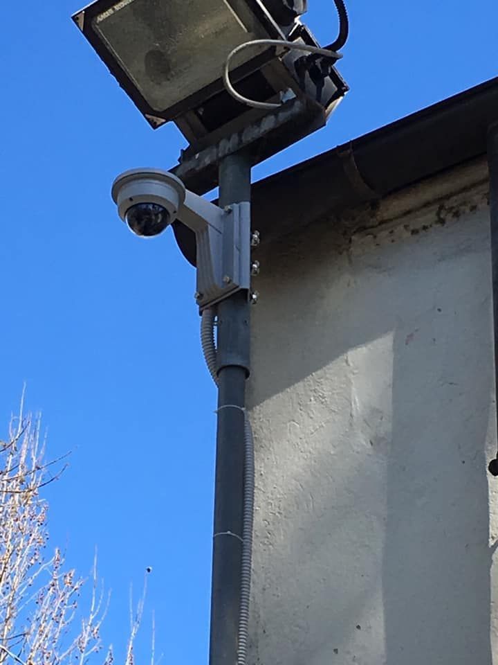 Telecamera di sicurezza e faretto montati su un palo accanto a un edificio, contro un cielo azzurro.