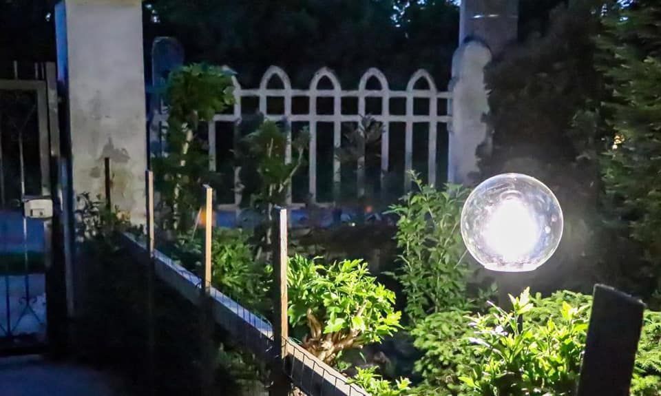 Una lampada da giardino a forma di sfera illumina un giardino verde, delimitato da una recinzione scura di fronte e da una recinzione decorativa bianca sul retro.