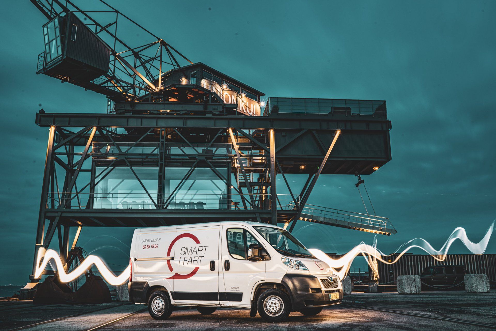 En hvid varevogn er parkeret foran en stor kran om natten.