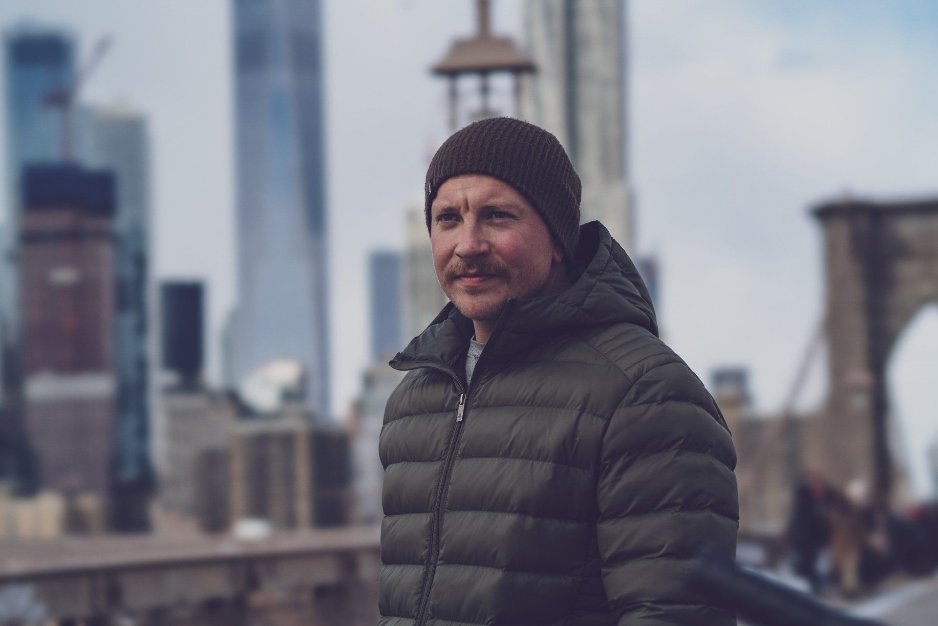 En mand iført hat og jakke står foran byens skyline.