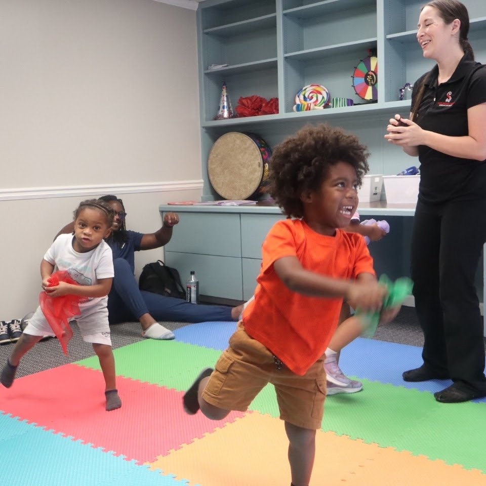 Children playing in a colorful room with an adult. The children hold colorful streamers and weights, the adult smiles.