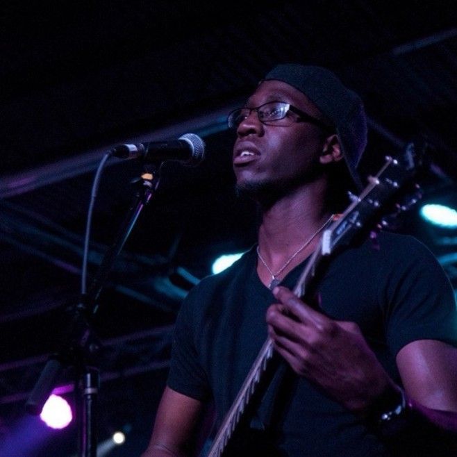 Man playing guitar and singing into a microphone on stage, under blue and purple lights.