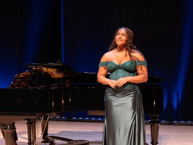 Woman in green dress on stage next to piano, looking upward, smiling. Blue backdrop.