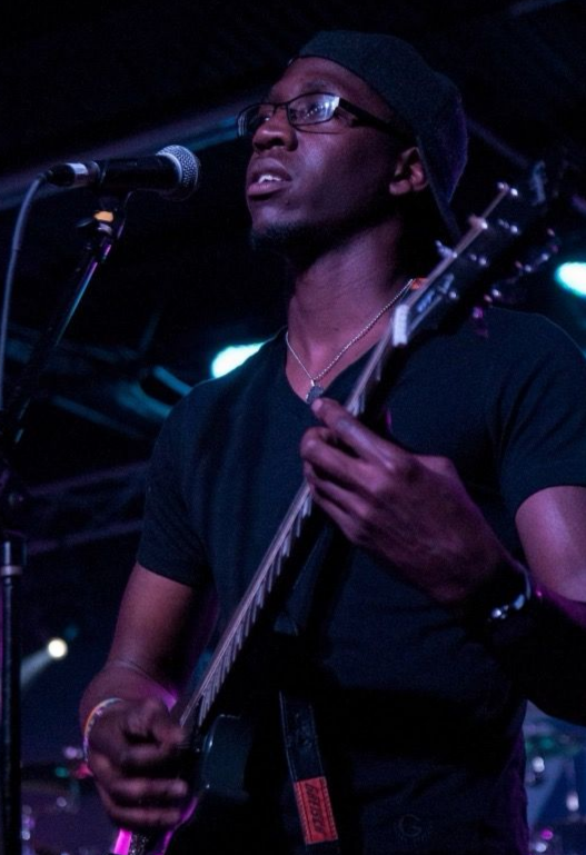 Man playing a guitar and singing into a microphone on stage, wearing a black shirt and a beanie.