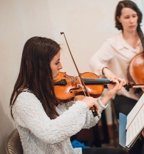 A woman with long hair is smiling and wearing a black jacket and a necklace.