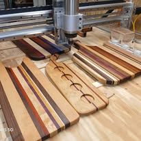 Wooden cutting boards in various designs on a workbench with a CNC machine in the background.