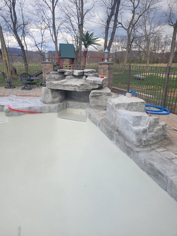 Pool with waterfall feature and gray stonework. Overcast day with a metal fence in the background.