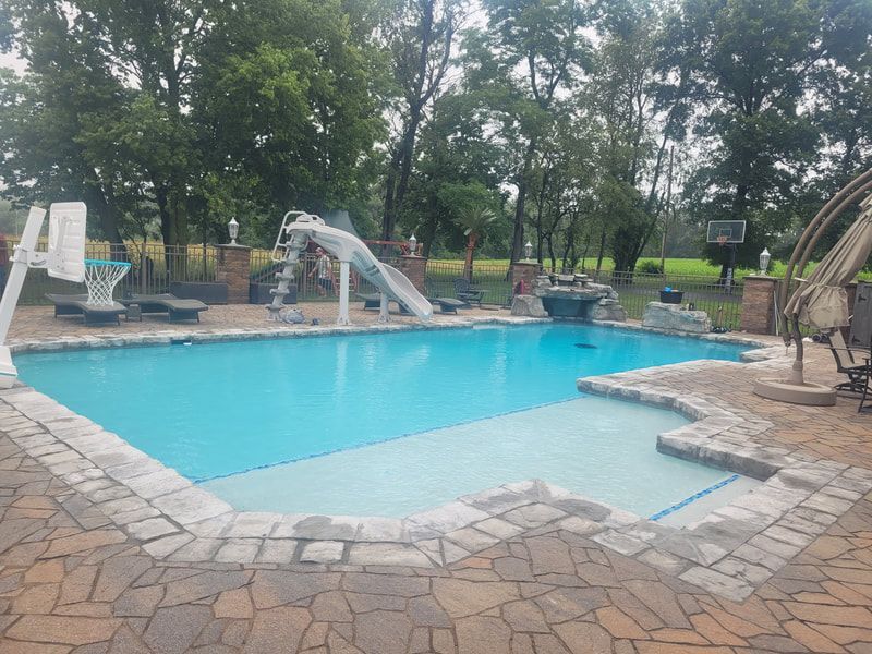 Swimming pool with slide, basketball hoop, and stone patio, surrounded by trees.