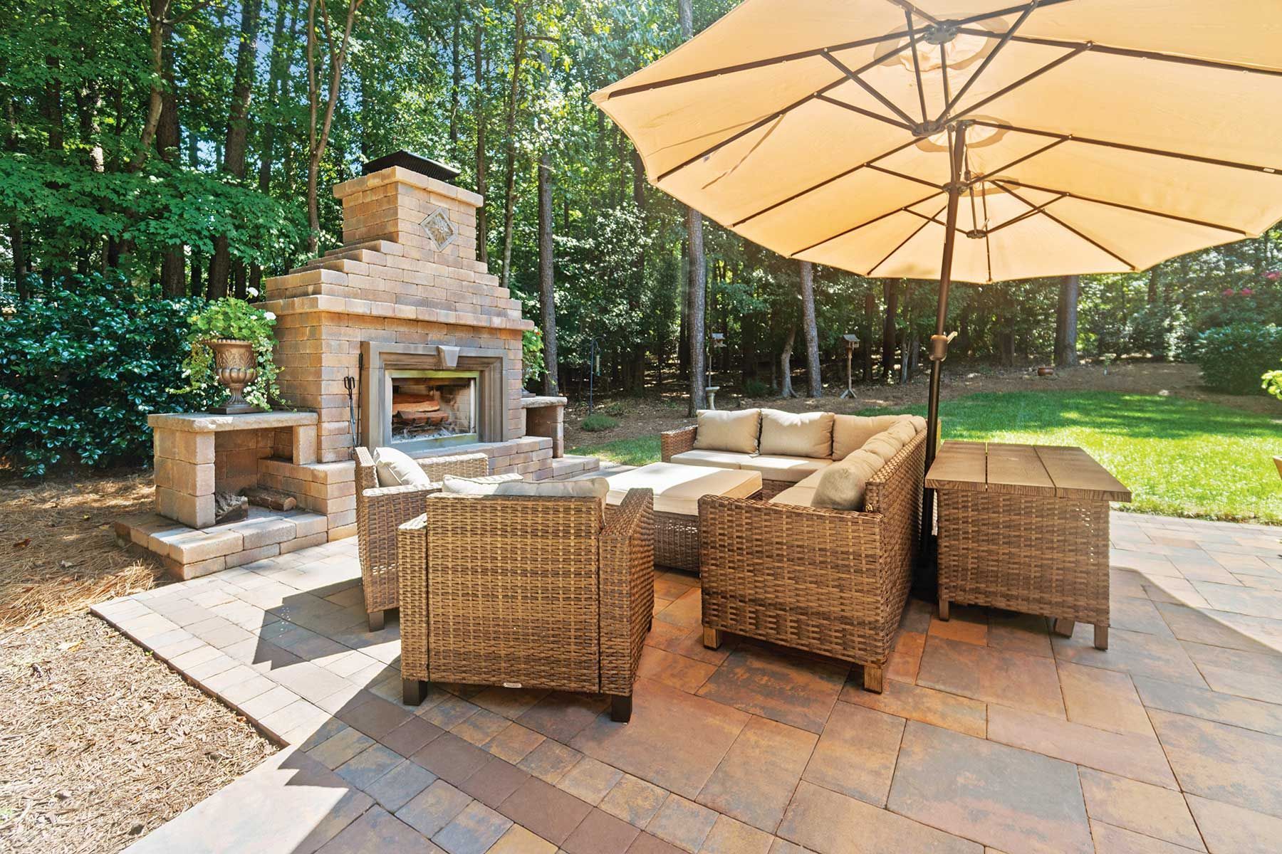 Outdoor patio with fireplace, seating area, and umbrella surrounded by trees.