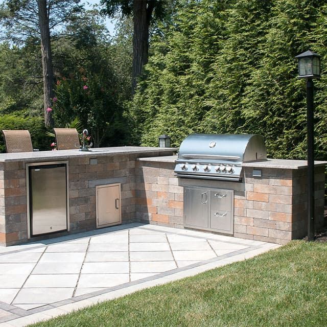 Outdoor kitchen with built-in grill, refrigerator, and storage cabinets on a stone patio, with two chairs.