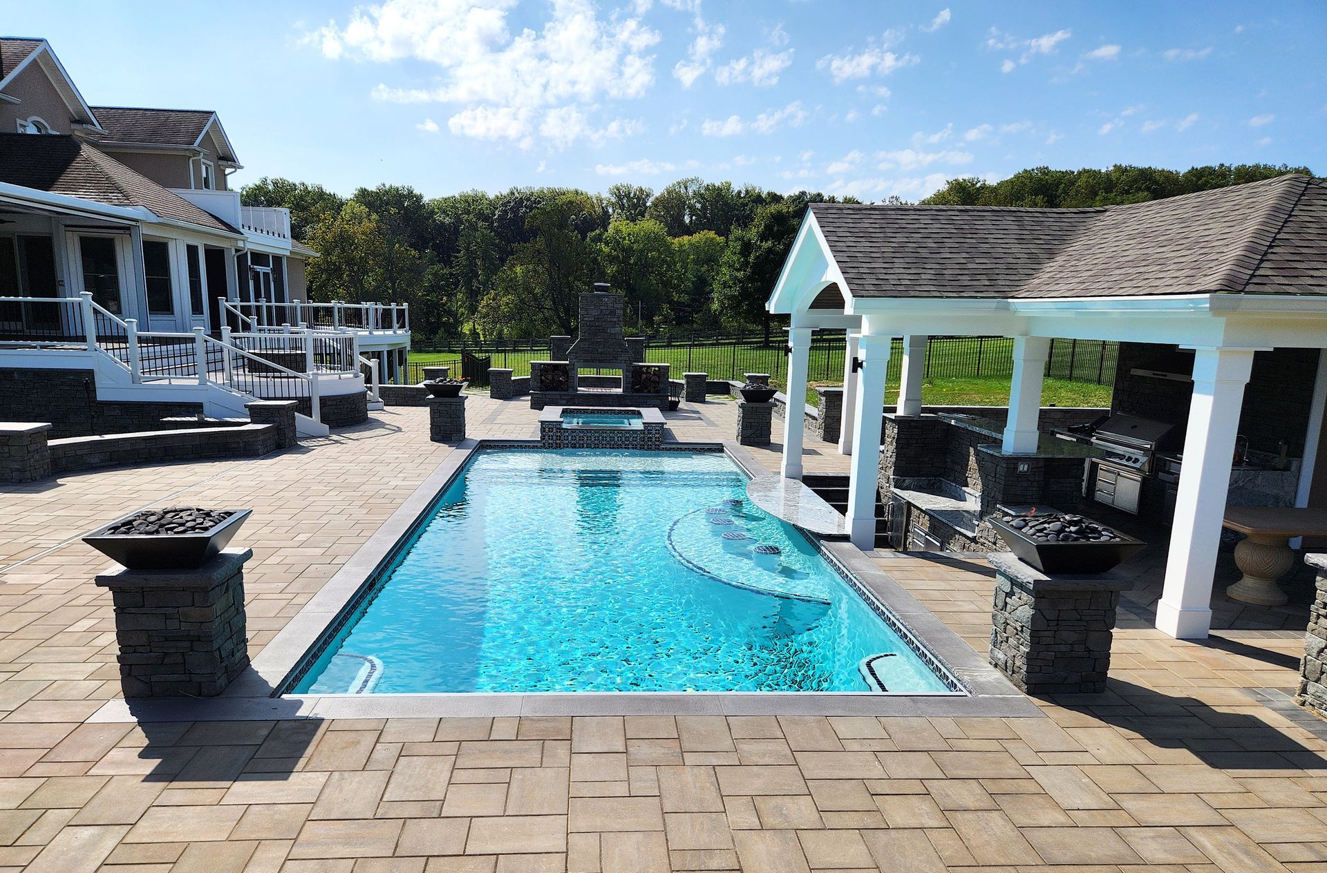 Rectangular blue swimming pool in a paved backyard with a gazebo, fire pits, and a large house.