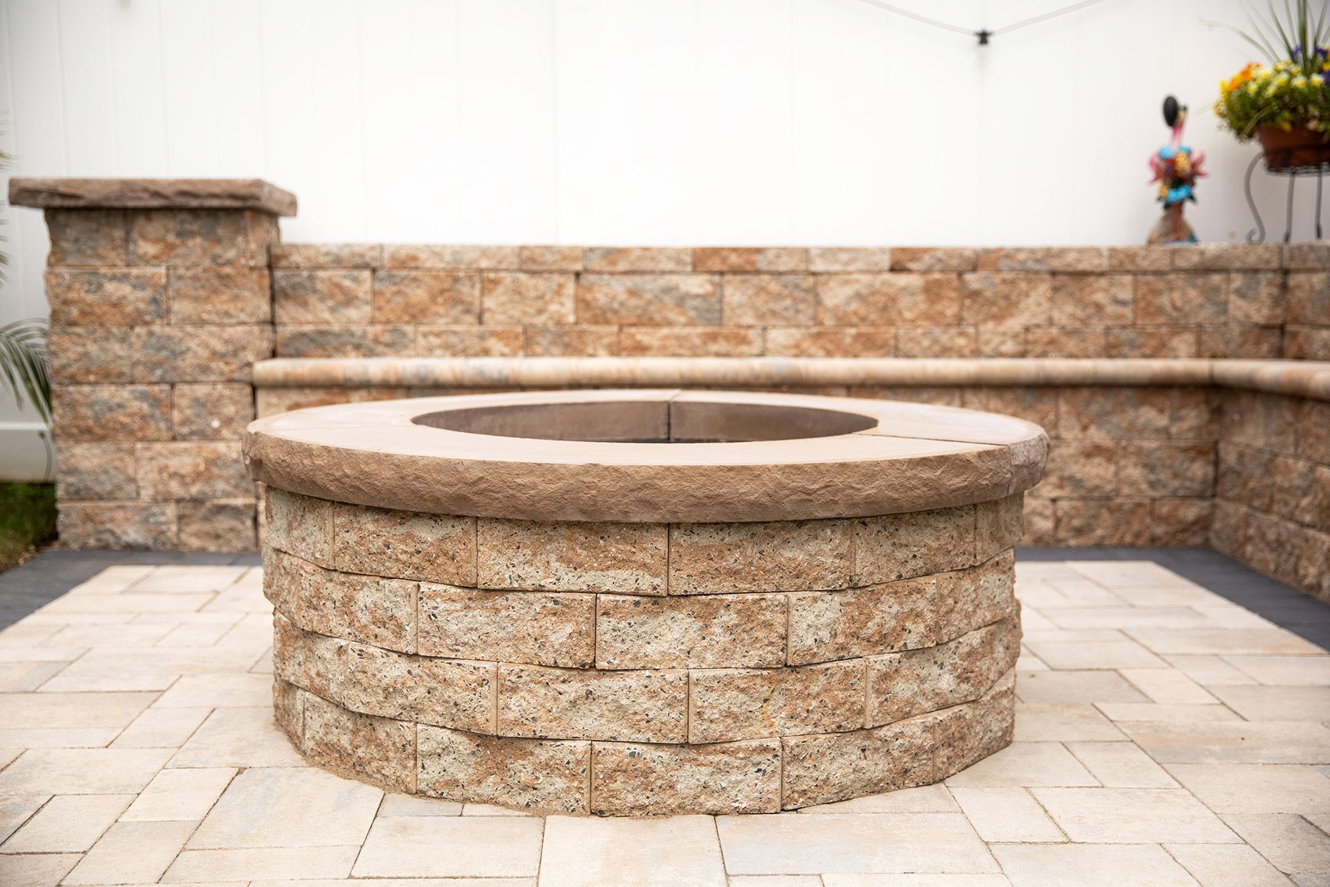 A round brick fire pit on a stone patio with a built-in bench in a backyard.