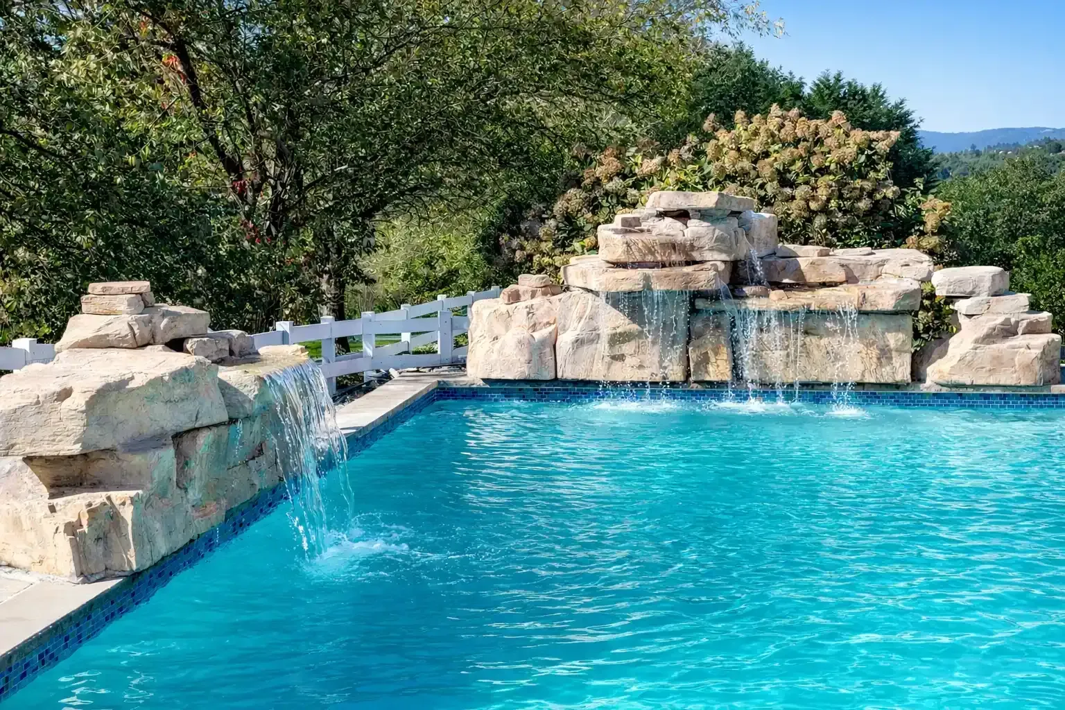 Blue pool with stone waterfall feature, white fence, and greenery in background.