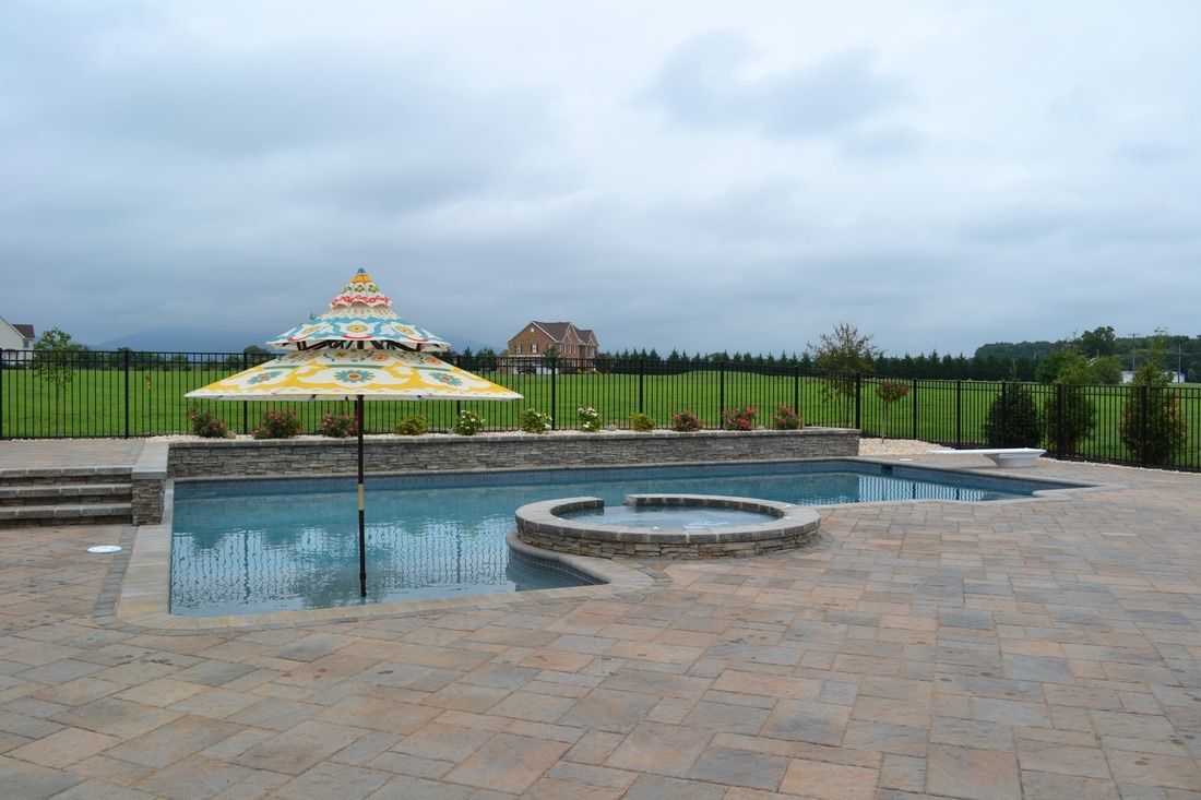Backyard pool with umbrella, stone patio, and black fence against a cloudy sky.