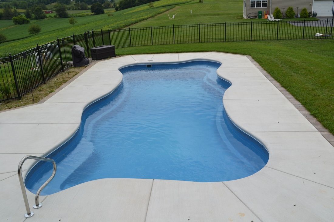 Swimming pool with blue water and concrete deck, set in a green backyard with a fence.