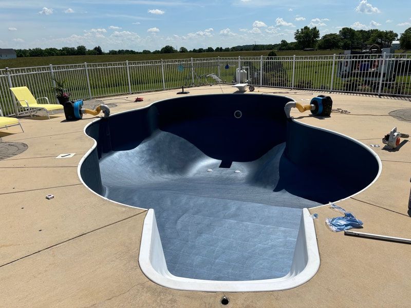 Empty kidney-shaped swimming pool with blue interior and concrete surround, near a white fence, under a blue sky.