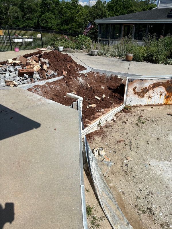Dirt and debris on a concrete patio next to a rusty metal frame, likely a pool. Buildings and foliage in background.