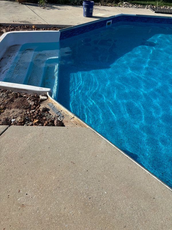 A partially filled swimming pool with blue water and concrete steps. Concrete edging surrounds it.