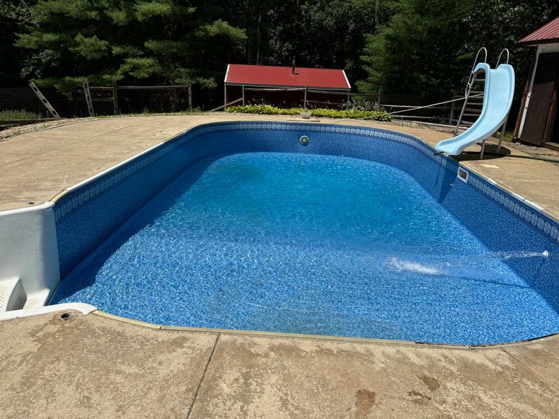 Blue-tiled swimming pool with a water slide, surrounded by concrete, with a red-roofed structure and trees in the background.