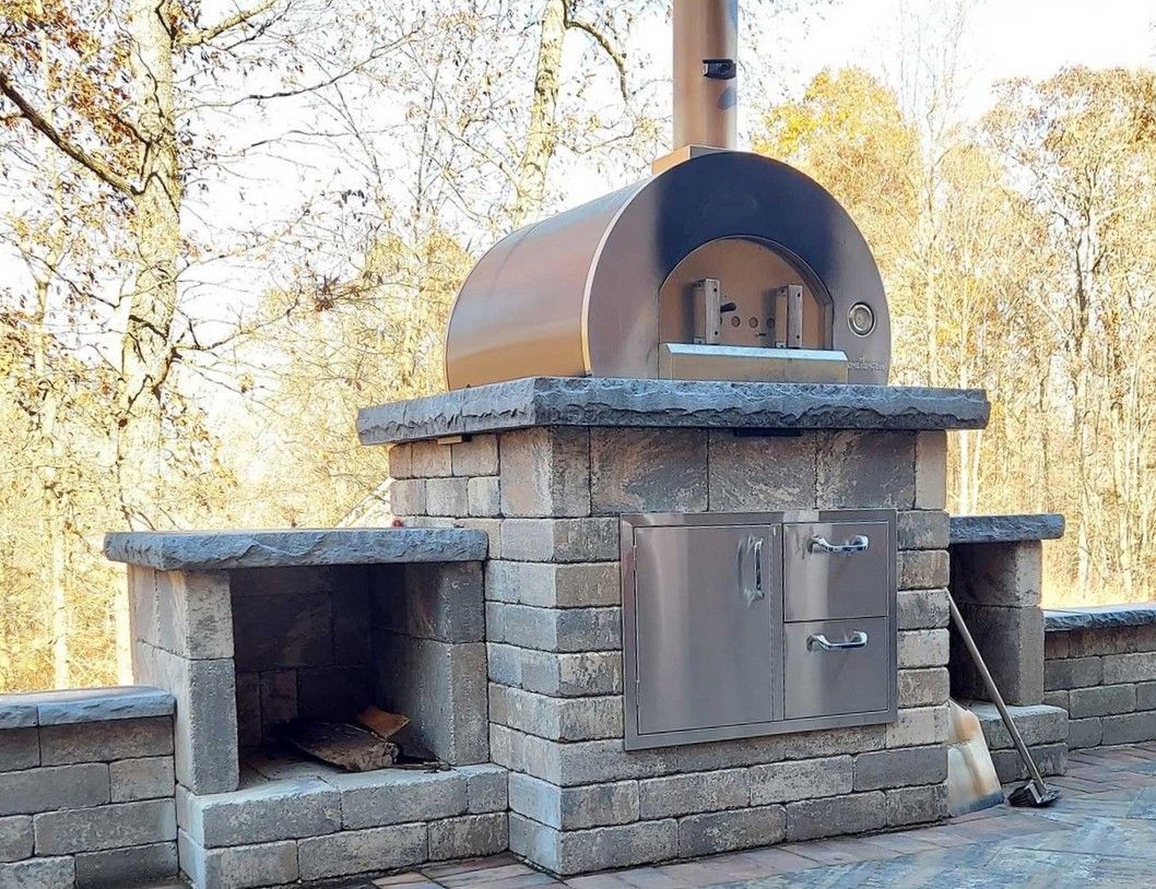 Outdoor brick pizza oven with stainless steel door and chimney, on a stone patio.