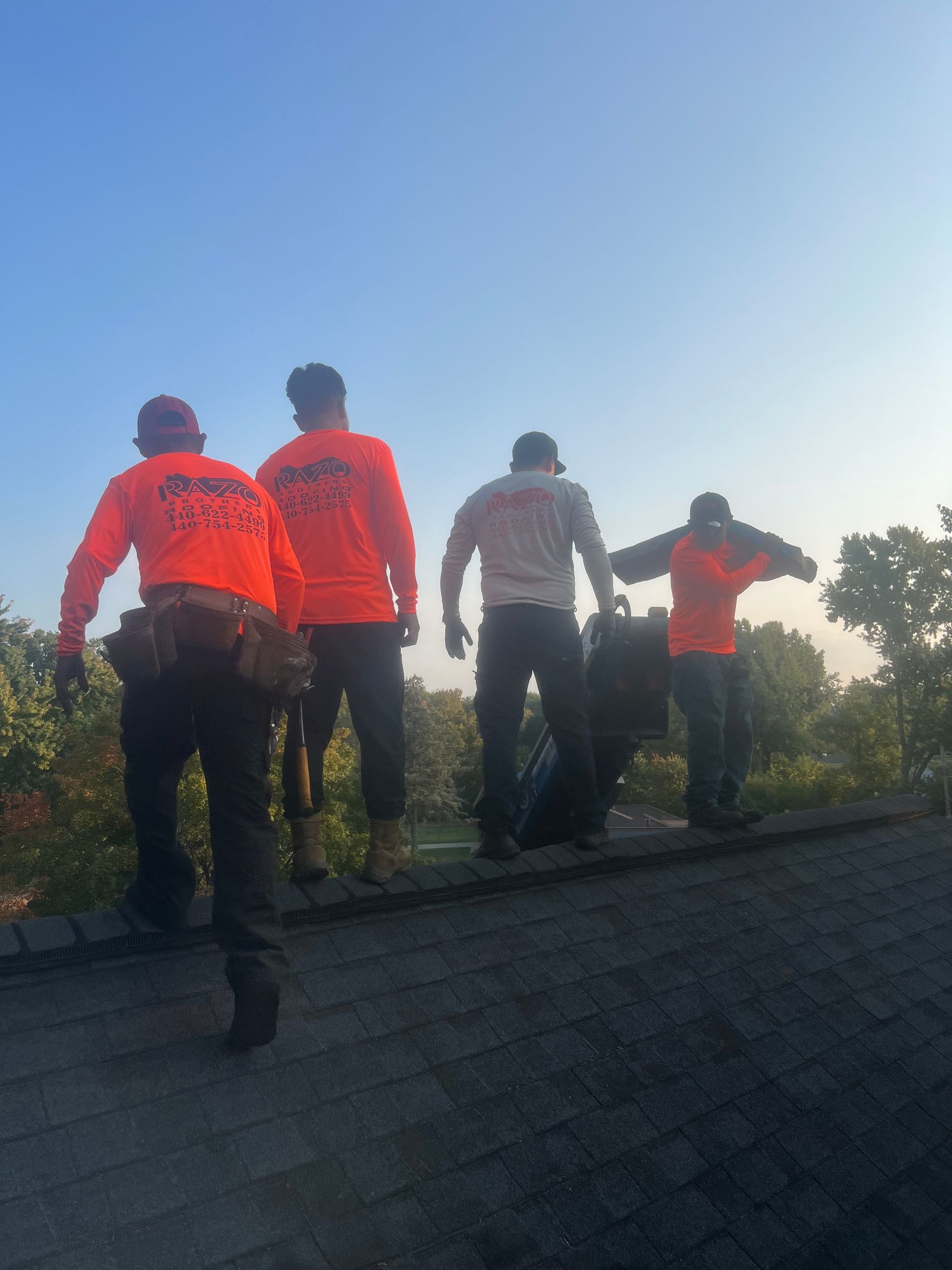 Four roofers, backs to camera, walk across a dark gray rooftop. One carries a black bag. Bright, early sunlight.