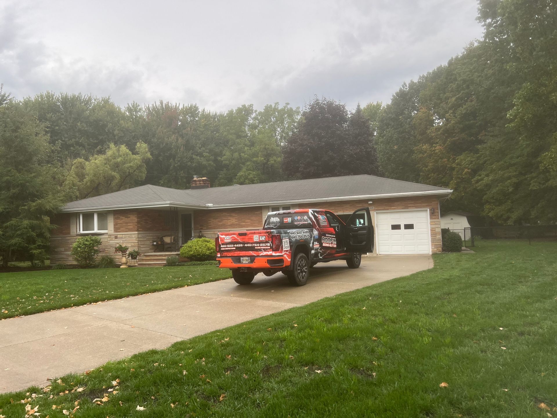 Red truck parked in front of a brick house with a white garage door, trees in the background, overcast sky.