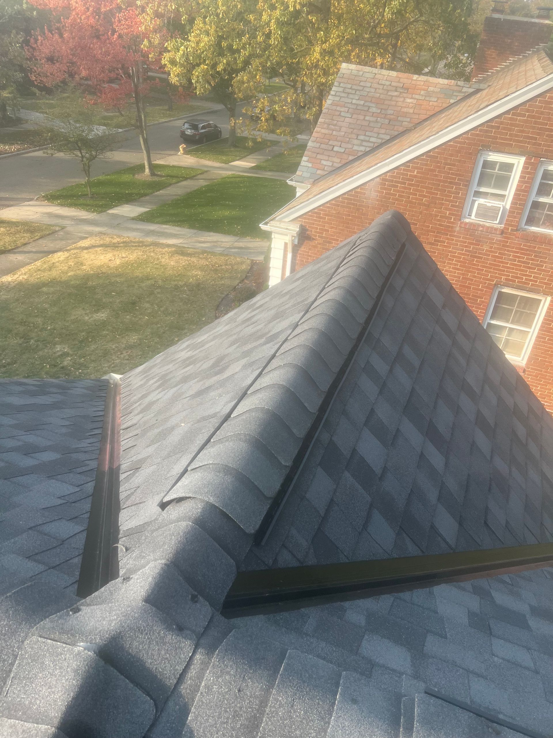 View of a house roof with grey asphalt shingles. Nearby is another brick home and autumn trees.