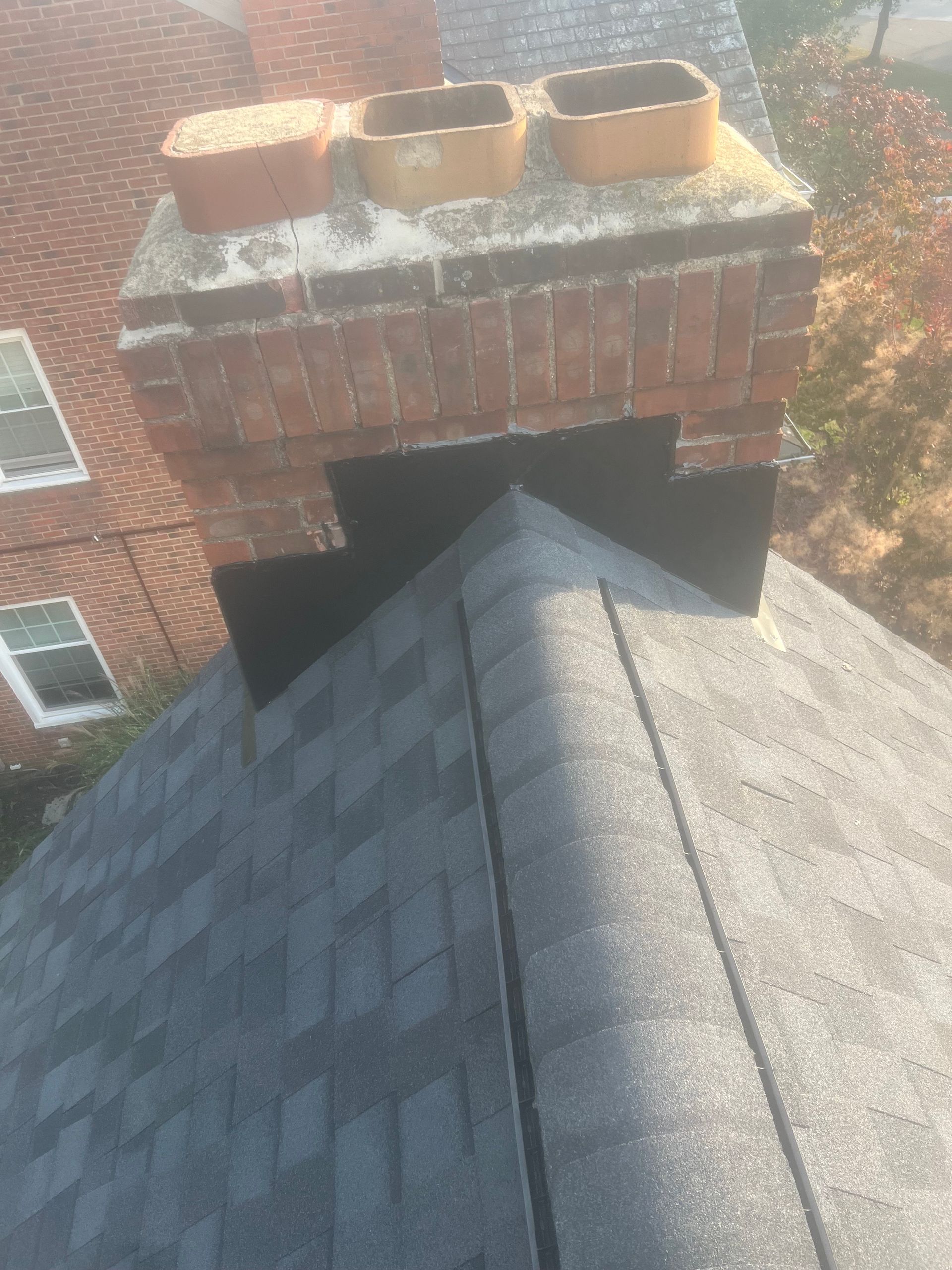 Rooftop view of a chimney with three flues and dark shingles. Flashings surround the chimney base.