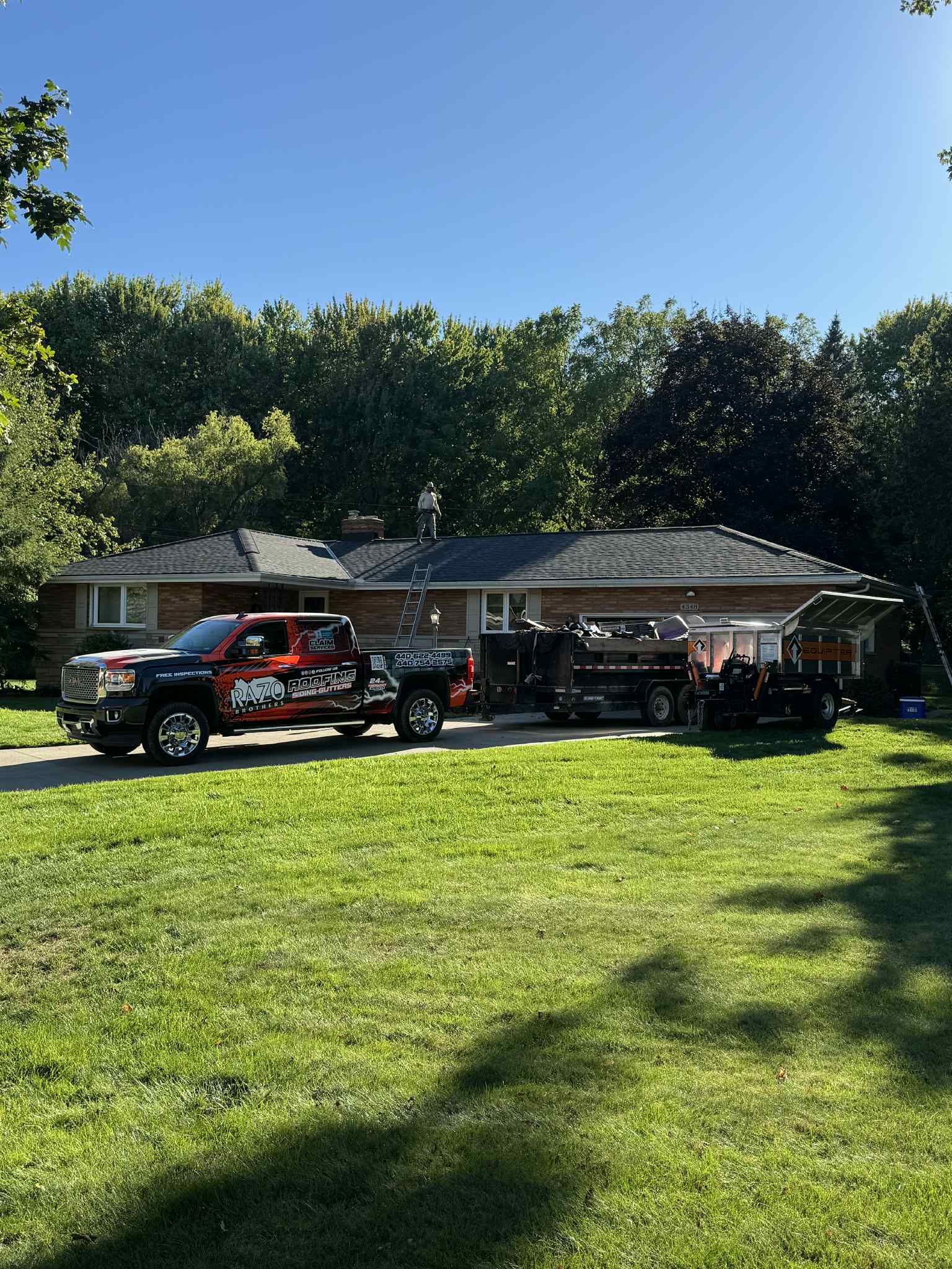 A truck and trailer parked in front of a house, possibly for a roofing job, on a sunny day.