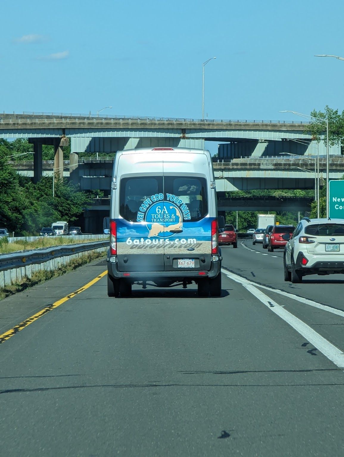 White van with advertisement on the highway, driving between other vehicles under a bridge on a sunny day.
