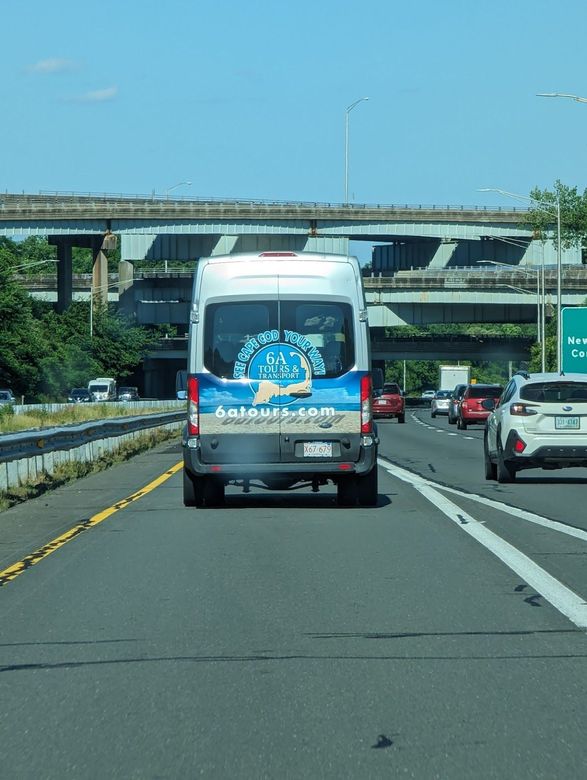 A white and blue commercial van drives on a highway under an overpass, accompanied by other vehicles.