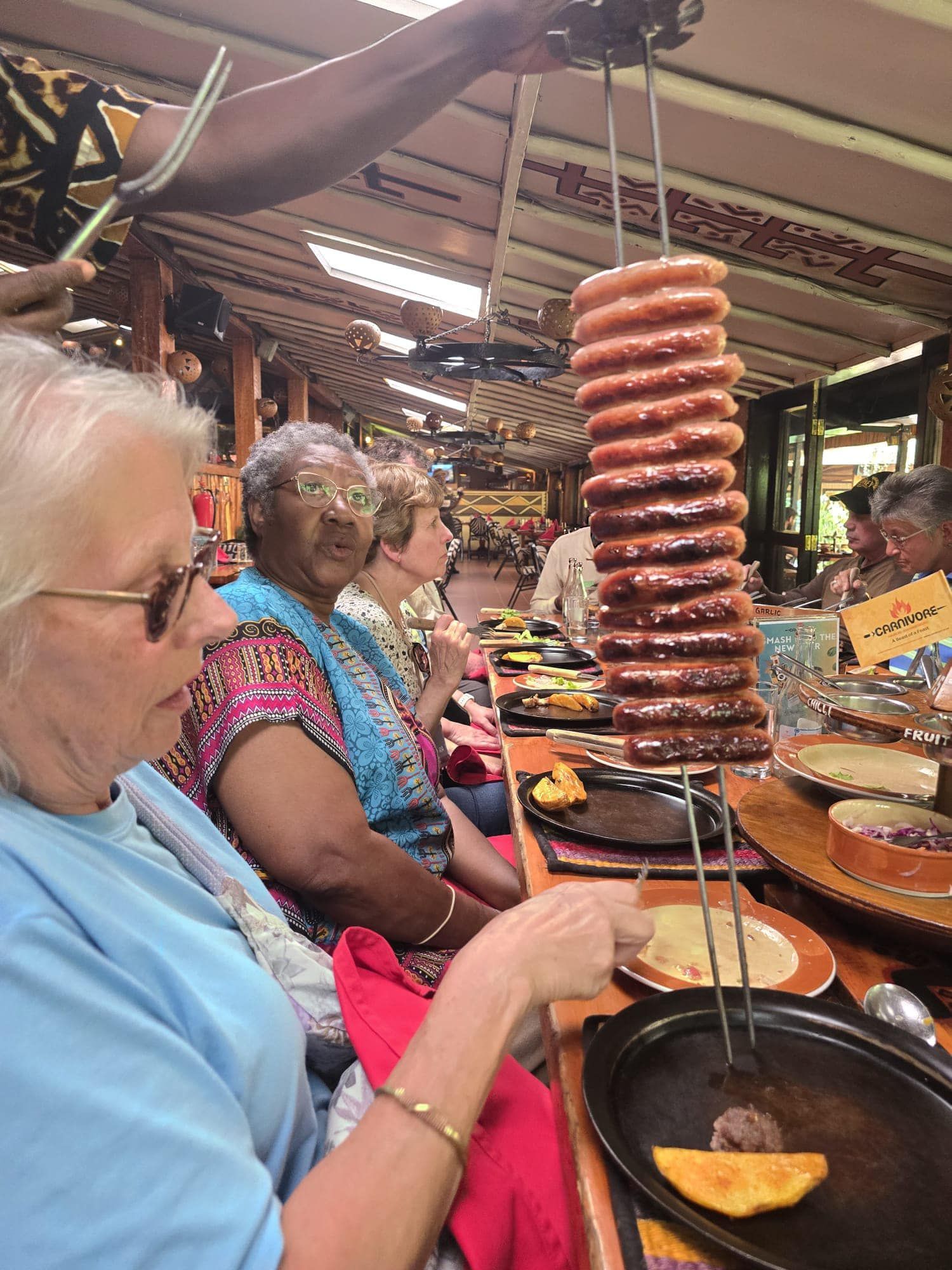 People dining at a restaurant with hanging grilled sausages. A woman uses tongs.
