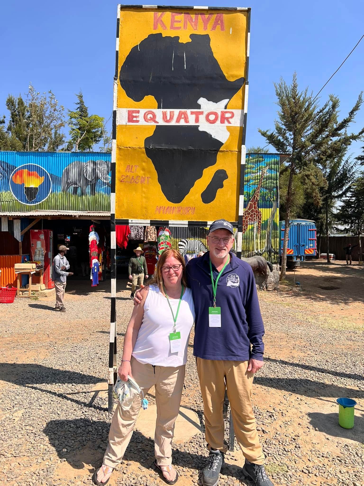 Two people pose near a Kenya Equator sign, with shop in the background.