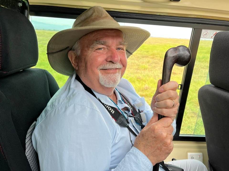 Man in safari hat smiles, holding cane, seated in vehicle, grassy background.