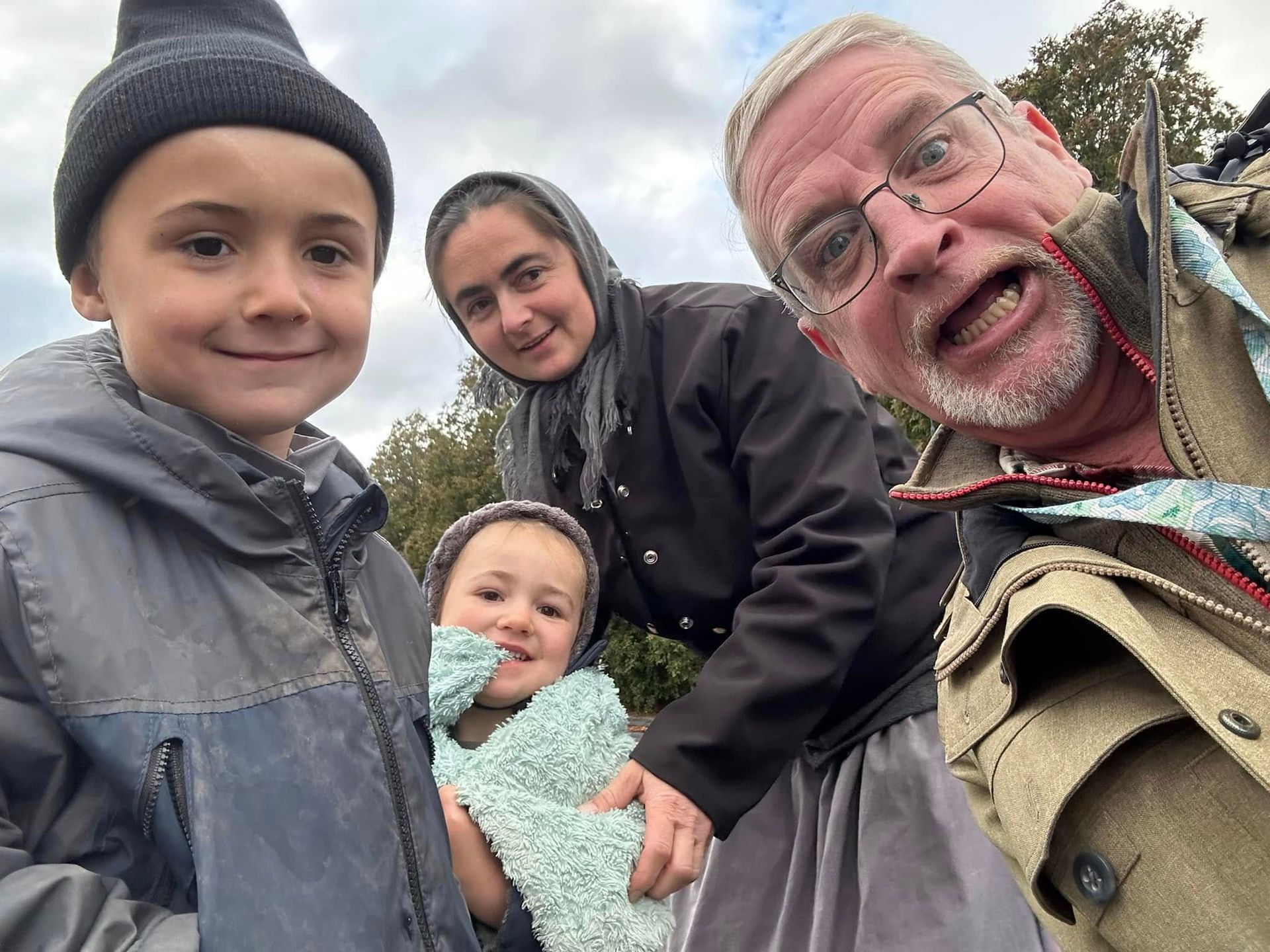 Group selfie outdoors: four people, one man grinning, a woman with head covering, and two children smiling.